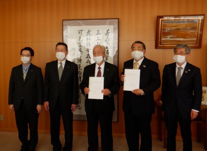 書道作品が壁に掛けられている前で複数の男性のうち2人の男性が書類を手に持っている写真