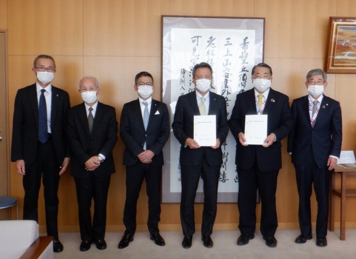 書道作品が掛けられている壁の前で手を前で組んでいる男性や書類を手に持っている男性などが並んでいる写真