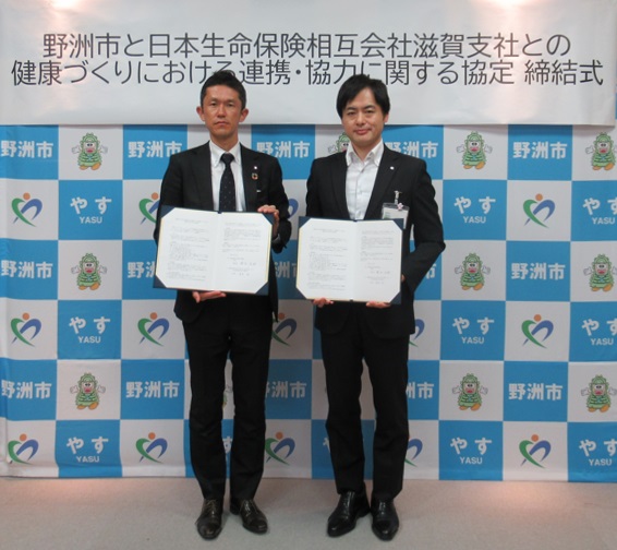 「日本生命保険相互会社滋賀支社と健康づくりにおける連携・協力に関する協定 締結式」の垂れ幕の前で二人の男性が一緒に書類を持っている写真