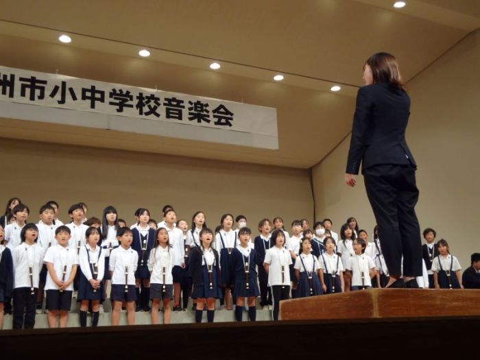 野洲市小中学校音楽会と書かれた横断幕を背に、指揮者の前に並んで制服姿の生徒たちが合唱している写真。