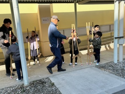 竹馬の乗り方指導の様子。5名の小学生が竹馬に乗ろうとしている写真。大人2名が指導している。
