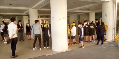 学生帽をかぶった小学生たちと、制服を着た中学生たちが、校舎の入口に集まっている様子。