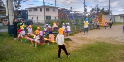 学生帽をかぶった幼稚園生たちが、シーソーなどのカラフルな遊具で遊んでいる。