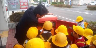 学生帽をかぶった子供たちが、赤い郵便ポストの前に集まっている写真。大人がポストの扉を開け、使い方を説明している。
