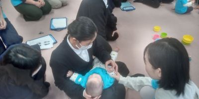床に座った制服姿の子どもたちが赤ちゃんと関わっている写真。赤ちゃんは子どもの膝の上で抱っこされており、大人が赤ちゃんの手を持って補助している。