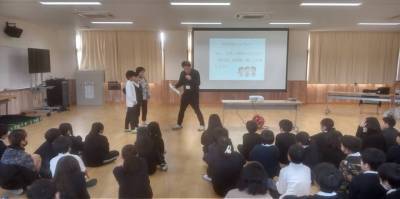 ロールプレイの場面。屋内で、プロジェクターを使って発表を行っている。二人の小学生とマイクを持った所長が前に立ち、他の子どもたちは座ってそれを見守っている。