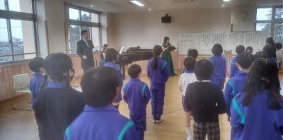オペラ歌手から指導を受ける様子。前方に教師とピアノがあり、子供たちは立って歌っている。
