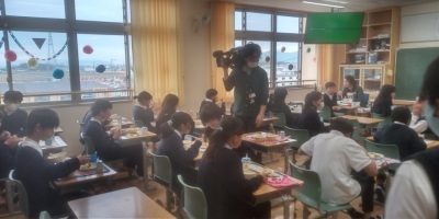 教室で席に座って給食を食べる小学生たちと、その様子を撮影するカメラマンの写真。