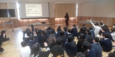 がん教育出前授業。カーテンを閉めた部屋で保健師がスクリーンを使って講義を行い、生徒たちが座って聞いている。何人かは手を挙げている。