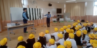 交通安全教室。教室で警察官が小学生に交通安全を指導している様子。学生帽をかぶった小学生が前方に座り、警察官がホワイトボードを使って説明している。
