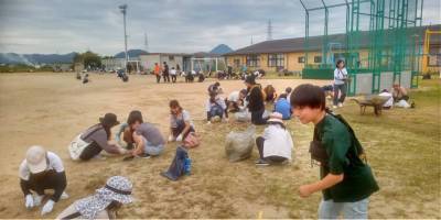 親子除草作業。校庭の草地に沿って並んでしゃがみ、除草をしている大人と小学生たち。みな軍手をつけて作業している。
