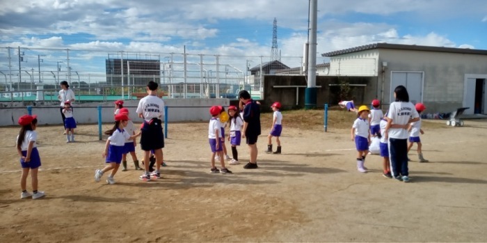 校庭の鉄棒の近くで、走ったり立ったりしている小学生たちと中学生たち。小学生は体操服で赤い帽子をかぶっており、中学生は体操服を着ている。