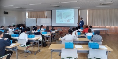 教室で授業を受ける小学生たち。生徒たちは机に座り、前方のスクリーンに映し出された資料を見ている。講師が前に立ち説明を行っている。