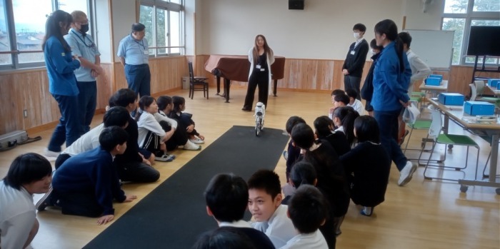 教室で小学生がロボットを囲んで見学している写真。教師やスタッフも周囲に立ち、ロボットの動きを観察している。
