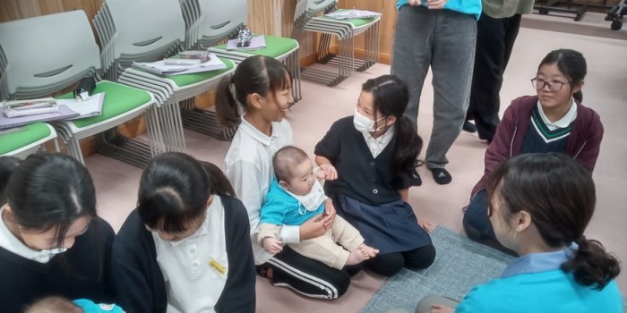 赤ちゃんとのふれあい。小学生が床に座り、赤ちゃんを抱っこしている写真。隣にはハグベビーのスタッフが付き添い、笑顔で会話している。