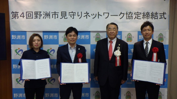 「第4回野洲市見守りネットワーク協定締結式」と書かれた横断幕の前で、4人中3人が書類を手にし立っている写真