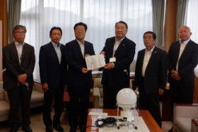 机に置かれている投光器をバックに複数の男性のうち2人の男性が書類のようなものを持ち並んでいる写真