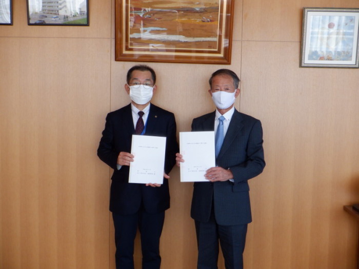 マスクを着用したスーツ姿の男性2人が胸の高さで協定書のようなものを手に持ち並んでいる写真