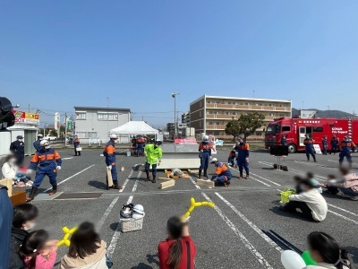 屋外で数名の消防団員が演習のようなことをしていてる様子を複数の人が周りで座って見ている写真