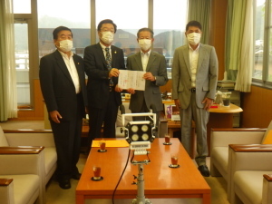 会議室のような所でスーツとマスクを着用した4人の男性のうち2人の男性が協定書のようなものを手に持って並んでいる写真