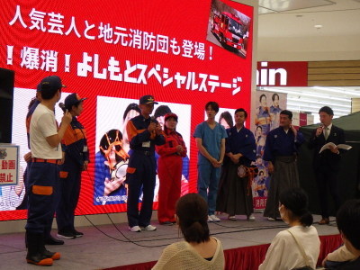 消防団員や袴やスクラブ、スーツなど様々な服装の男女がステージ上に立っている様子を複数の人が見ている写真