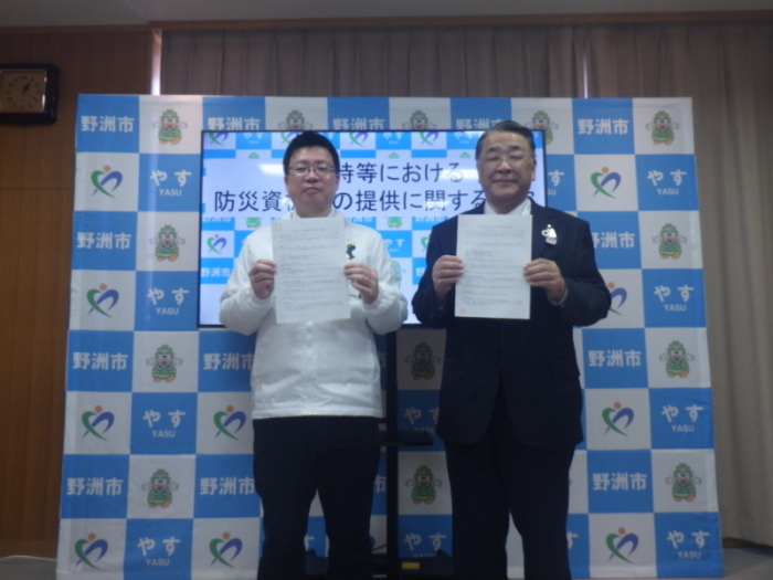 モニターが置かれた部屋で書類を手に持った2人の男性が並んでいる写真