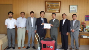 発電機が置かれている後ろにスーツを着用した複数の男性が立っており、真ん中の男性2人は協定書のようなものを手に持って並んでいる写真