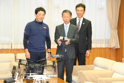 3人の男性のうちスーツを着た1人の男性がドローンを操縦するコントローラーを手に持ちもう1人の男性に動かし方の説明を聞いている写真