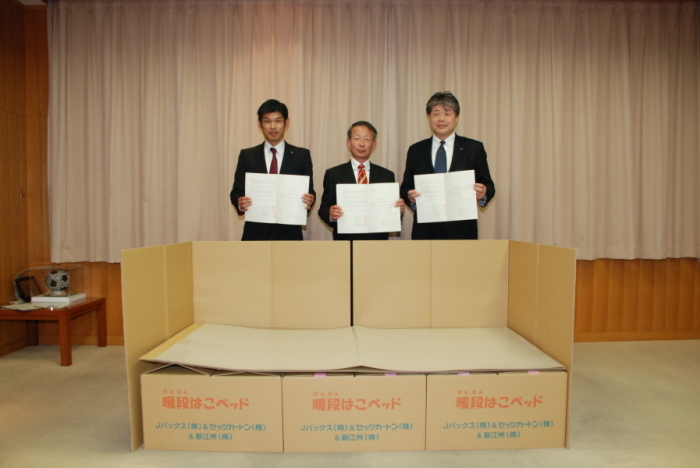 段ボールでできたベッドの後ろに協定書のようなものを手にしたスーツ姿の男性が3人並んでいる写真