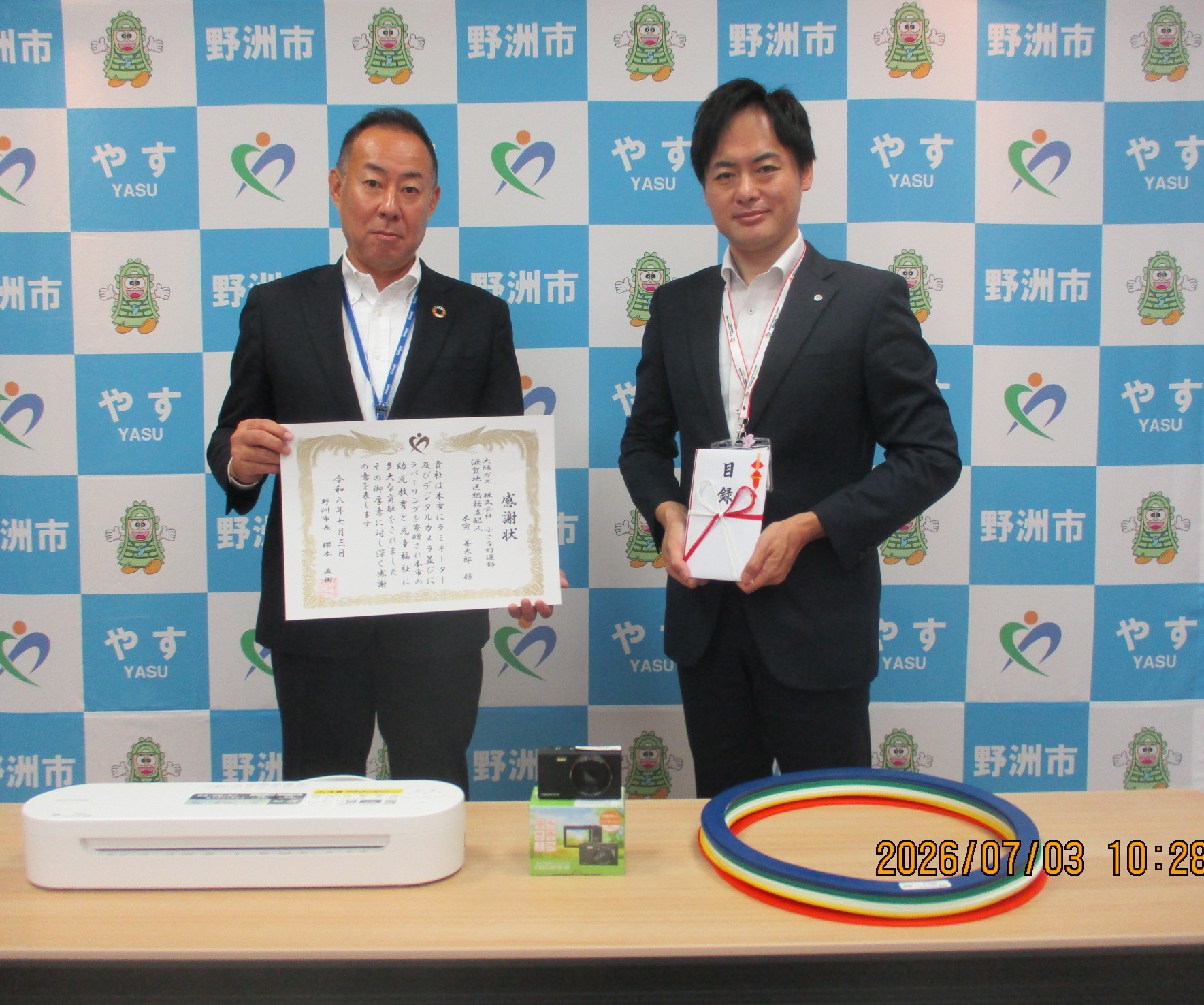 50代の男性2名のうち、右側の男性は目録と書かれた封筒を持ち、左側の男性は賞状を持っている写真