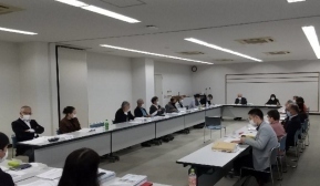 広い会議室に四角く配置された長机に沢山の職員がついて座り、資料を見ながら会議している様子の写真