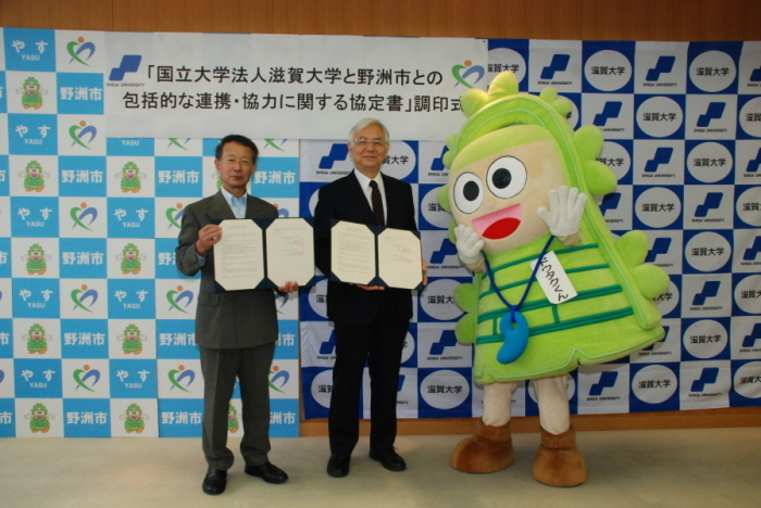 書状を持っている2人の男性と市のマスコットの着ぐるみが立っている写真
