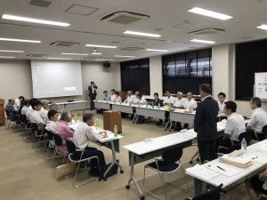 広い会議室に白い長机が四角く囲むように配置され、沢山の職員が座り、会議をしている様子の写真