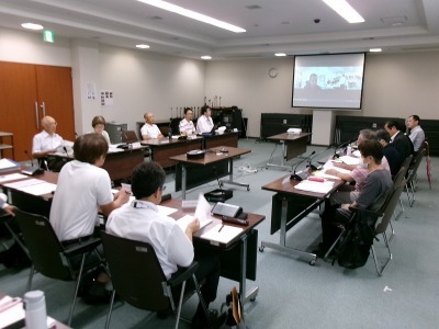 会議室でスクリーンに映し出された人と椅子に座っている参加者が会議を行っている様子の写真