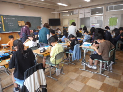 教室内で席に座って裁縫をする児童たちに4人の女性たちがそれぞれアドバイスをしている写真