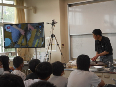 特別教室内で男性が魚をさばきながらその様子をモニターに映して児童たちに解説をしている写真