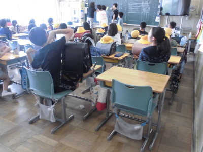 教室内で机にランドセルや黄色い帽子が置かれた机に着席したり黒板の前に立ったりしている男女の児童たちの写真