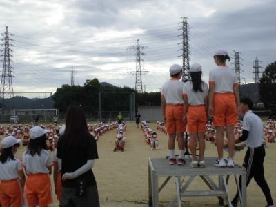 運動場の朝礼台に立つ子供たちとそれを体育座りでみる子供たちの写真