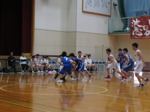 ユニフォームを着用した複数の生徒が試合を行っている様子を周りで複数の生徒が椅子に座って見ている写真